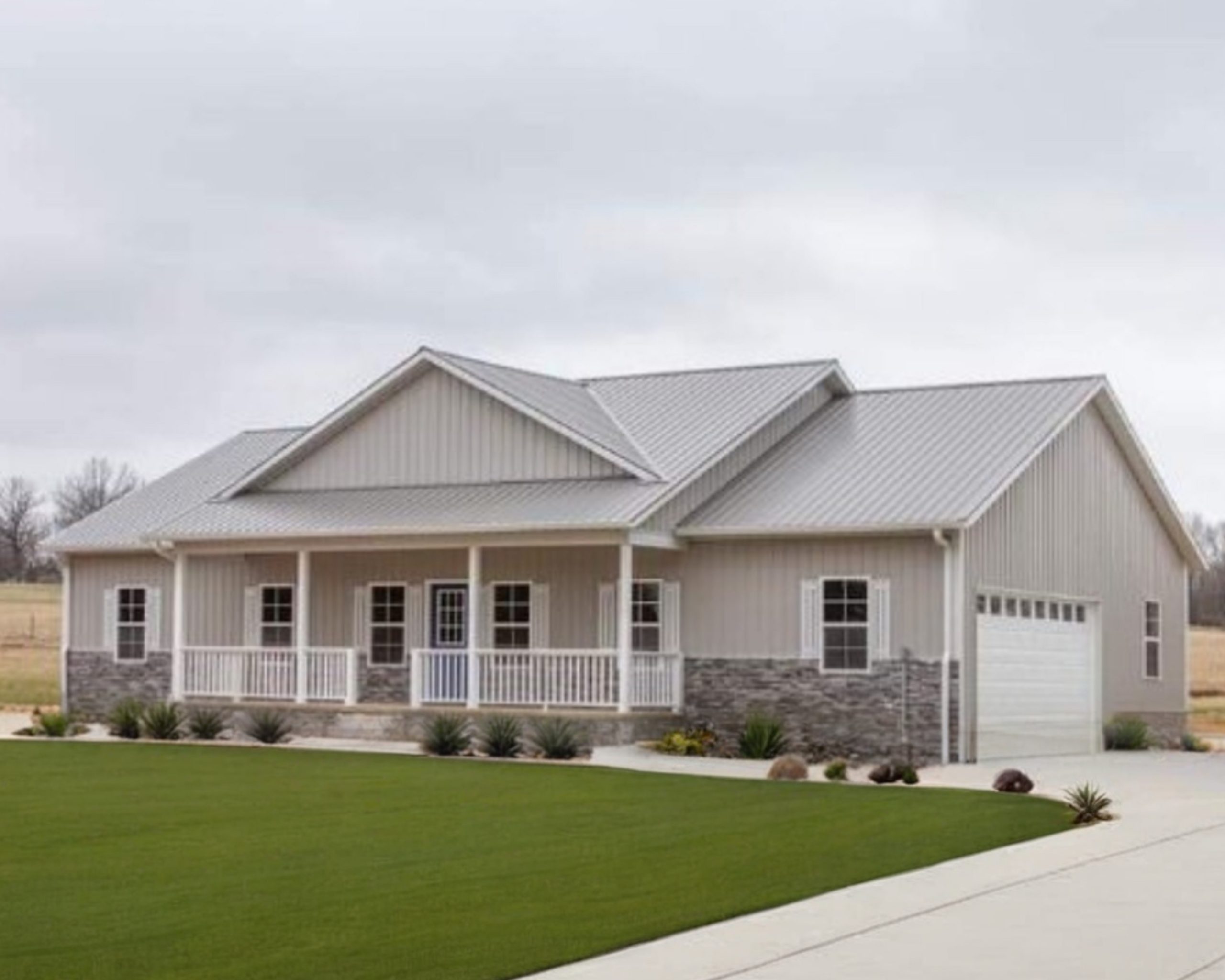The Cornerstone (Plan #15A) — 4 bed/2 bath, 1820 sq ft wood-framed house kit, exterior view with light tan board-and-batten siding, gray standing-seam metal roof, covered front porch with white columns, stone wainscoting, and attached 2-car garage